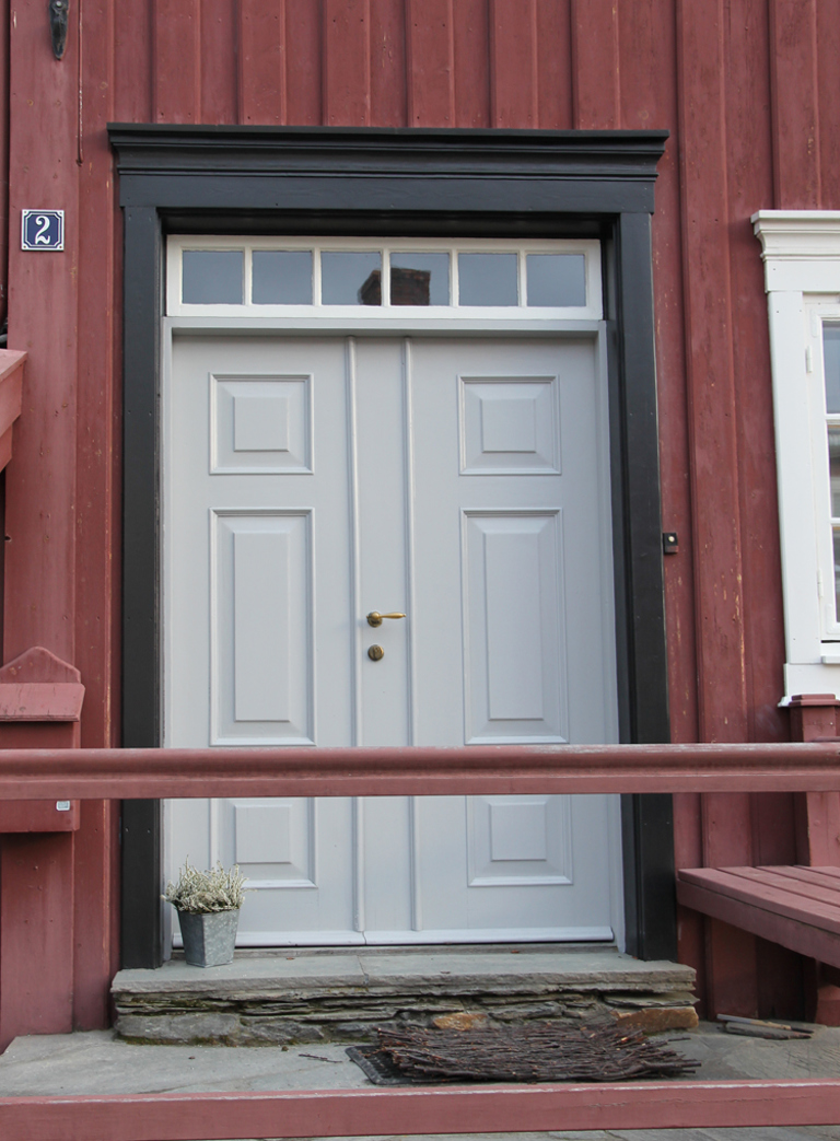 lys grå umbra dør med mørkebrun ubrent umbra omramming mot rød kledning. Hvitt vindu. Røros. Foto: Christel Wigen Grøndahl, Bygg og Bevar